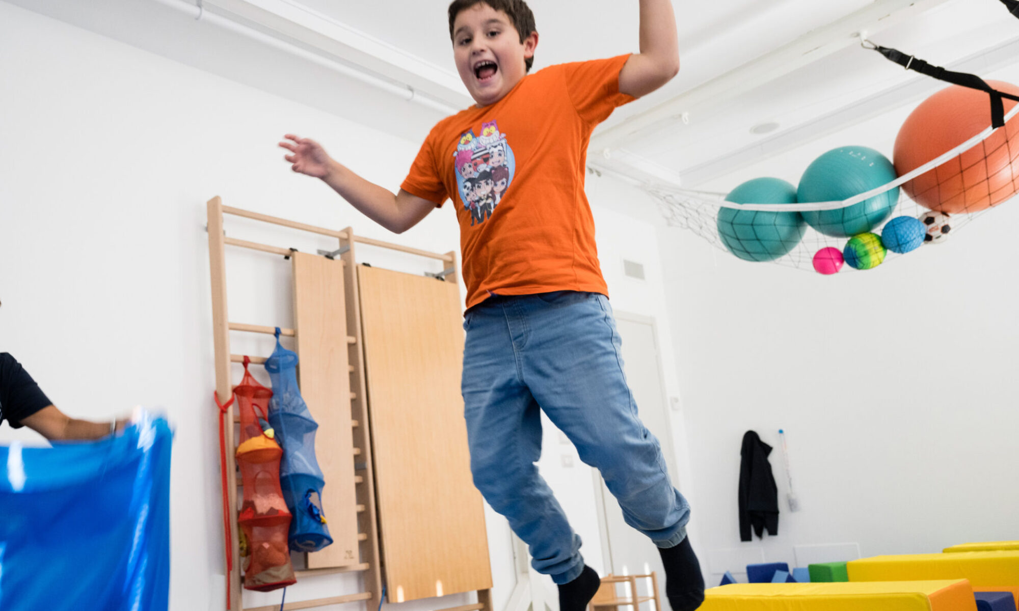 Bambino che salta con entusiasmo su materassi colorati in una sala attrezzata per attività motorie, circondato da palloni e giochi educativi.