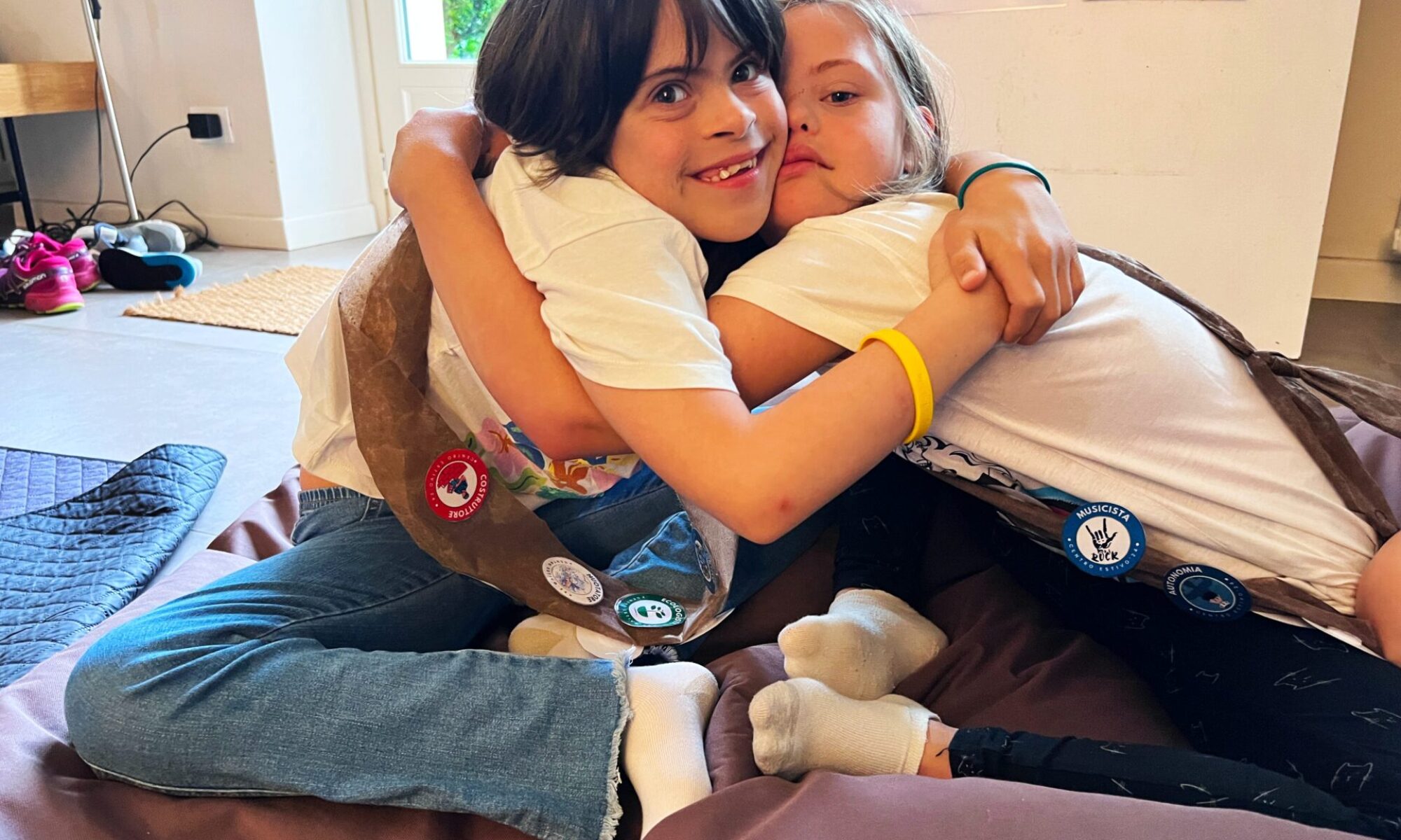 Due bambine sedute su un cuscino che si abbracciano, indossando fasce con distintivi e sorridendo in un ambiente accogliente e luminoso.