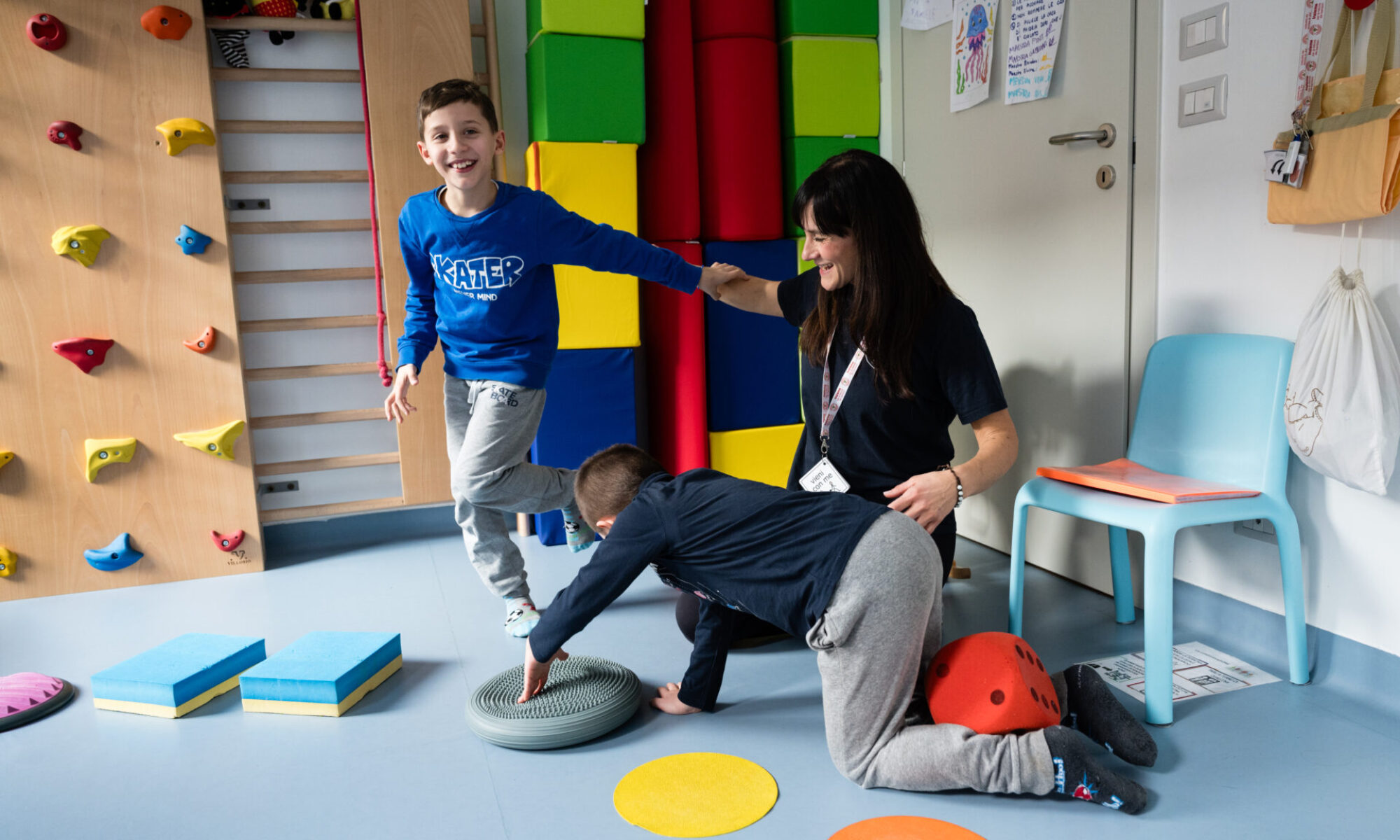Bambino che sorride durante un'attività motoria su tappetino, accompagnato da un’operatrice in un ambiente colorato con attrezzi per psicomotricità.