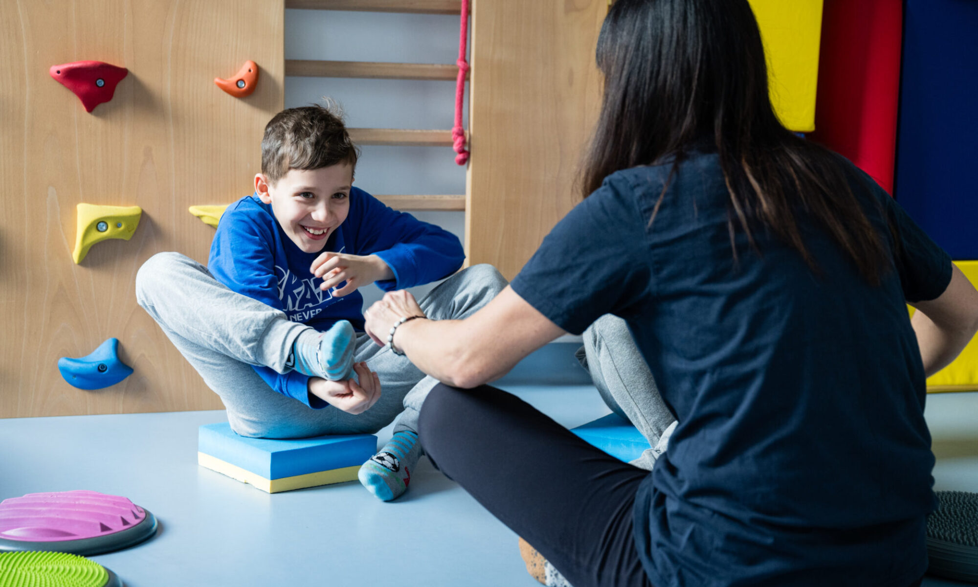 Bambino che sorride durante un'attività motoria su tappetino, accompagnato da un’operatrice in un ambiente colorato con attrezzi per psicomotricità.